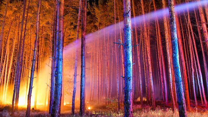 24.08.2018, Brandenburg, Klausdorf: Hell erleuchtet ist ein brennender Wald nahe Klausdorf, der gerade versucht wird, mit einem Wasserstrahl aus einem Fahrzeuges der Feuerwehr zu löschen. Ein Feuer hat sich im Südwesten Brandenburgs auf einer großen Waldfläche ausgebreitet. Rund 300 Einsatzkräfte bekämpfen den Waldbrand südwestlich von Berlin. Die Behörden ließen drei Dörfer räumen. Foto: Patrick Pleul/dpa-Zentralbild/dpa +++ dpa-Bildfunk +++