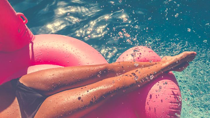 Woman floating on a pink inflatable in swimming pool. Tight crop showing only her legs and feet. She is tanned in turquoise water with water splashing onto her.
