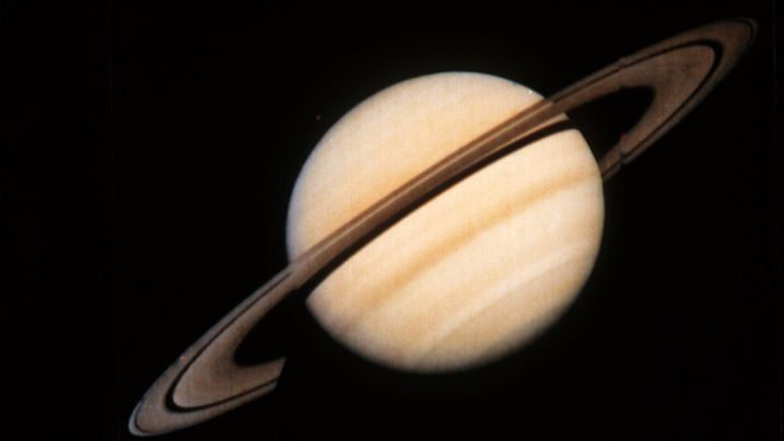 Der Saturn mit seinen Ringen, von der Sonde Voyager 1 der US-Raumfahrtbehörde NASA am 18. Oktober 1980 übermittelt. Der Planet Saturn ist der zweitgrößte Planat im Sonnensystem. Er besteht aus Wasserstoff und Helium und hat von allen Planeten die geringste Dichte und größte Abplattung. Aus Bahnvermessungen der Raumsonden Pioneer 11 und Voyager 1 und 2 wird ein fester und eisenhaltiger Kern vermutet. Saturn besitzt eine dichte Athmosphäre mit streifiger Wolkenstruktur, in der enorme Wirbelstürme mit Windgeschwindigkeiten bis zu 1800 km/h auftreten. Das auffällige Ringsystem besteht aus Tausenden von Einzelringen sehr geringer Dicke. 23 Monde des Saturn sind bekannt (Mimas, Enceladus, Tethys, Dione, Rhea, Titan, Hyperion und Iapetus) und weitere werden vermutet.