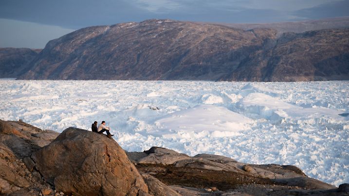 ARCHIV - 16.08.2019, Grönland, Helheim Glacier: Zwei Studenten der New York University, die hier an einer Forschungsexkursion teilnehmen, sitzen auf einem Felsen mit Blick auf den Helheim-Gletscher. Am höchsten Punkt des grönländischen Eisschildes hat es am Wochenende zum ersten Mal seit Beginn der Wetteraufzeichnungen geregnet. (zu dpa "Erstmals Regen auf grönländischem Eisschild in 3200 Metern Höhe") Foto: Felipe Dana/AP/dpa +++ dpa-Bildfunk +++