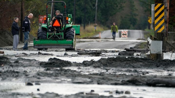 A tractor operator talks with people standing near a mud- and debris-covered road near Everson, Wash., Monday, Nov. 29, 2021. Localized flooding was expected Monday in Washington state from another in a series of rainstorms, but conditions do not appear to be as severe as when extreme weather hit the region earlier in November. (AP Photo/Elaine Thompson)