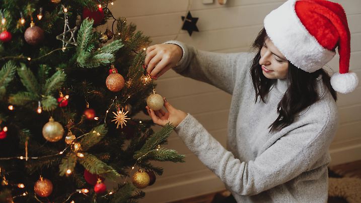 Happy young woman in santa hat decorating modern christmas tree with golden glitter bauble in festive decorated room with lights. Winter holiday preparations. Merry Christmas