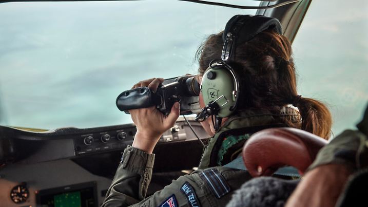 In this photo provided by the New Zealand Defense Force, a crew member from an Orion aircraft looks out the window as it flies over an area of Tonga that has heavy ash fall from a volcanic eruption in an area of Tonga, Monday, Jan. 17, 2022. Thick ash on an airport runway was delaying aid deliveries to the Pacific island nation of Tonga, where significant damage was being reported days after a huge undersea volcanic eruption and tsunami. (CPL Vanessa Parker/NZDF via AP)