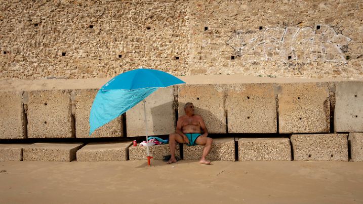 13.06.2022, Spanien, Cadiz: Ein Mann sitzt auf einem Wellenbrecher und schützt sich vor der Sonne unter seinem Sonnenschirm an einem Strand. Große Teile Spaniens werden in dieser Woche von einer für Juni ungewohnt heftigen Hitzewelle heimgesucht. Die höchsten Temperaturen wurden für Andalusien im Süden des Landes vorhergesagt. Foto: Emilio Morenatti/AP/dpa +++ dpa-Bildfunk +++
