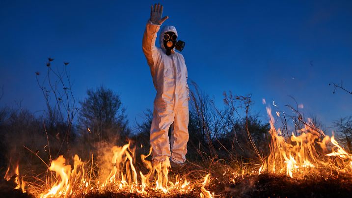 Fireman ecologist showing stop sign while working in field with wildfire at night. Man in protective radiation suit and gas mask near burning grass with smoke. Natural disaster concept.