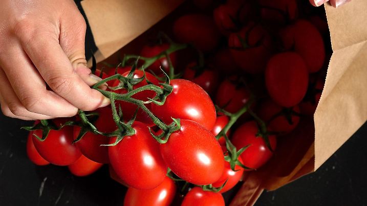 Detail on hands getting small red cherry tomatoes with green vines from brown paper bag, over dark black marble board.