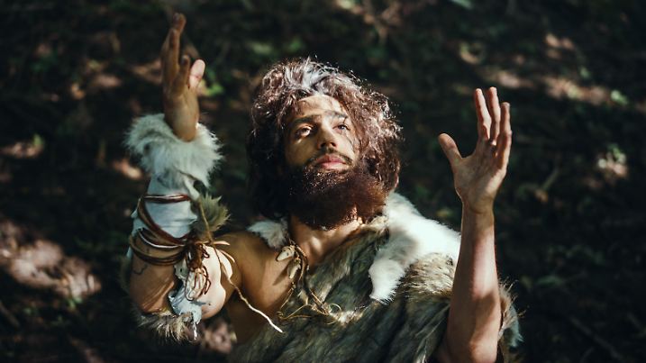 Portrait of Primeval Caveman Wearing Animal Skin Raises Hands to Heaven Looking at the Sun, Having Pagan Religious Experience. Prehistoric Neanderthal Believing and Praying to God. High Angle Shot