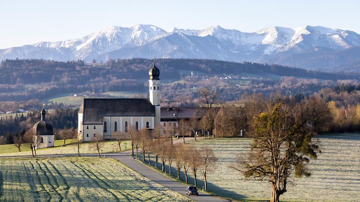10.04.2023, Bayern, Wilparting: Ein Auto fährt an der Wallfahrtskirche von Wilparting vorüber, während im Hintergrund die Alpen im Licht der morgendlichen Sonne zu sehen sind. Foto: Matthias Balk/dpa +++ dpa-Bildfunk +++