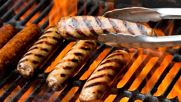 Bratwurst, hot dogs or polish sausage on a flaming charcoal grill. One brat is being presented in a pair of tongs in front of the others. All of them have beautiful appetizing grill marks as the flames kiss the meat sealing in the flavors.