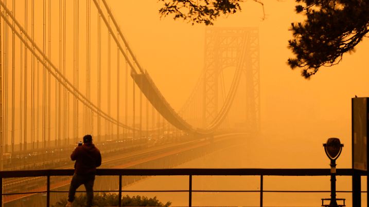 07.06.2023, USA, Englewood Cliffs: Ein Mann telefoniert, während er durch den Dunst vor der George Washington Bridge sieht. Waldbrände in Kanada hüllen den Nordosten der USA in Dunst und verfärbt den Himmel. Foto: Seth Wenig/AP/dpa +++ dpa-Bildfunk +++