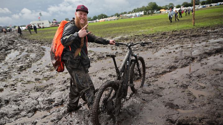 01.08.2023, Schleswig-Holstein, Wacken: Metal-Fan Lukas Litwin aus Bochum ist mit seinem E-Mountainbike auf dem schlammigen Festivalgelände unterwegs. Das Wacken Open-Air (WOA) vom 2. bis 5. August gilt als größtes Heavy-Metal-Festival der Welt und ist mit 85 000 Besucherinnen und Besuchern ausverkauft. Foto: Christian Charisius/dpa +++ dpa-Bildfunk +++