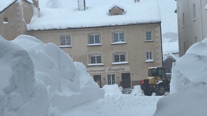 80 cm Neuschnee in dem Ort Simplon in der Schweiz an der italienischen Grenze. Ein Bagger schiebt die Schneemassen weg, um einen Weg zu schaffen. Im Hintergrund ist das Restaurant Simplon.