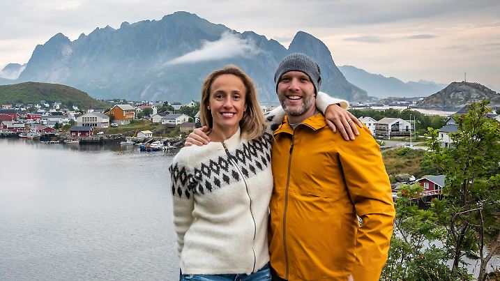 Couple visiting the fishing village of Reine in the Lofoten Islands with mountain range and cloudy sky in background during summer.
