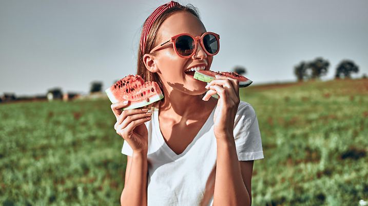 Sexy girl in red sunglasses holds a cut watermelon in her hands. Happy woman in trendy red sunglasses eat watermelon on nature