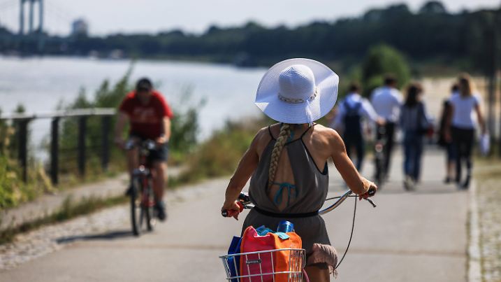 20.08.2024, Nordrhein-Westfalen, Köln: Eine Radfahrerin fährt mit Sonnenhut am Rheinufer entlang. Neben Sonnenschein und sommerlichen Temperaturen können sich die Menschen in Nordrhein-Westfalen in den kommenden Tagen auch auf etwas Abkühlung einstellen. Foto: Oliver Berg/dpa +++ dpa-Bildfunk +++