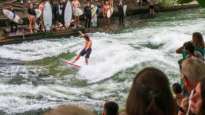 12.08.2024, Bayern, Landesweit: Zuschauer stehen bei hochsommerlichen Temperaturen am Ufer des Eisbach im Englischen Garten, der im Herzen der bayerischen Landeshauptstadt fließt, um den Surfern auf der künstlichen Wasserwelle zuzuschauen. Die Eisbachwelle ist zu jeder Jahreszeit ein wahrer Hotspot. Foto: Peter Kneffel/dpa +++ dpa-Bildfunk +++