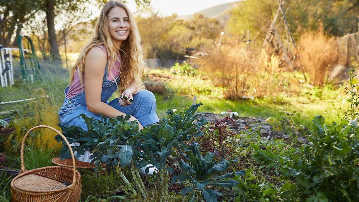 Portrait of a smiling young woman wearing dungarees kneeling in her organic vegetable garden examining her produce
