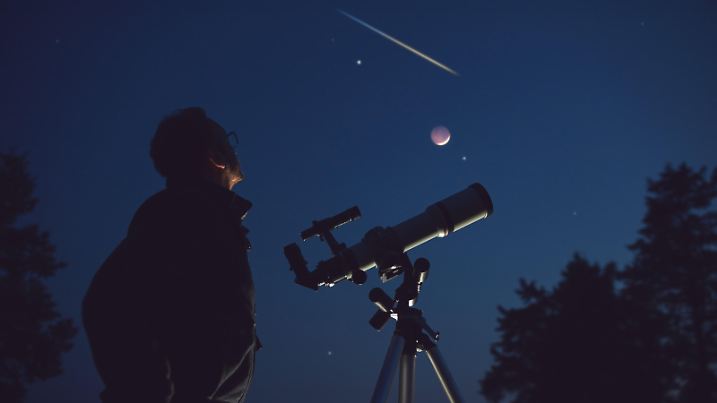 Silhouette of a man, telescope, stars, planets and shooting star under the night sky.