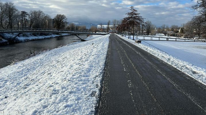 In Rosenheim hat es geschneit heute. Im Hintergrund sind die Alpen erkennbar, im Vordergrund eine geräumte Straße, rechts und links liegt Schnee.