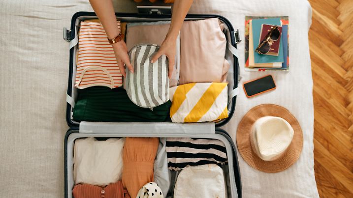 Shot from above of an anonymous woman packing things in her suitcase on the bed. She is holding and putting a grey and white stripped cosmetics bag in. There is her mobile phone, books and sunglasses on the bed.