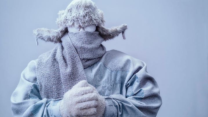 A frozen man with his hands clasped all bundled up in a fur trappers hat, scarf, parka and knit gloves, covered in snow and frost trying to stay warm on a very cold gray Winter's day.