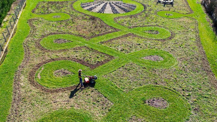 16.03.2025, Brandenburg, Jacobsdorf: Der Künstler Michael Uy bearbeitet mit einer Fräse den Boden auf seiner «Jakobswiese» im Landkreis Oder-Spree im östlichen Brandenburg (Luftaufnahme mit einer Drohne). Diese Wiese hatte Micheal Uy zusammen mit vielen fleißigen Helfern seit Anfang 2024 angelegt. Hunderte Arbeitsstunden hatte der Künstler in die Planung und Umsetzung der «Jakobswiese» investiert. Entstanden ist eine blühende Oase für die Bewohner des Ortes und natürlich für die vielen Insekten. Das Thema des blühenden Kunstwerkes ist «Der Lebensweg», denn diese Wiese liegt unmittelbar am Jakobsweg. Am Eingangsbereich ist eine Blumenrabatte in Form einer Jakobsmuschel zu sehen. Der Weg wurde in Form einer «Unalome» (Symbol für den Weg des Lebens) angelegt. Ein Pavillon und Sitzbänke laden zum Verweilen ein. Foto: Patrick Pleul/dpa - ACHTUNG: Nur zur redaktionellen Verwendung im Zusammenhang mit der Berichterstattung über das Projekt +++ dpa-Bildfunk +++