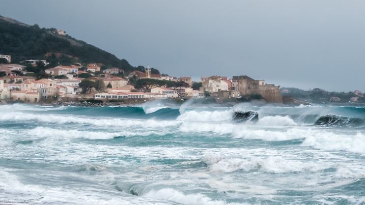 Raue See stürzt auf den Strand von Algajola in der Balagne-Region von Korsika mit Dorf und Zitadelle in der Ferne, 14.04.2022