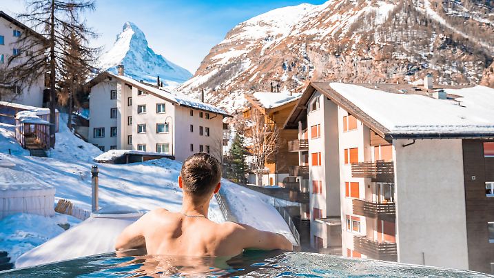 Tourist swimming in pool while looking at matterhorn mountains and houses. Rear view of man relaxing in hot tub. View of snow covered landscape in alps during winter.