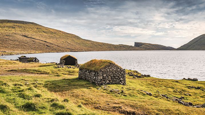 Grasdächer und Naturstein: traditionelle färöische Bootshäuser vor dem Sørvágsvatn-See unter blauem Sommerhimmel.