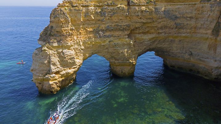A large boat passes under natural arches made of rocks in crystal clear water on the coast, aerial view, Praia da Marinh