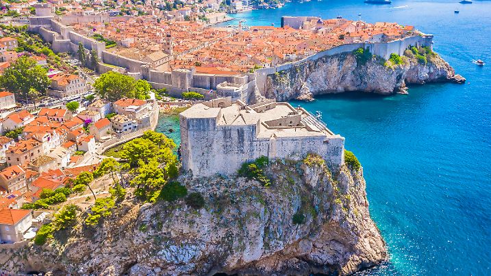 A stunning view from behind Fort Lovrijenac in Dubrovnik, Croatia, capturing the fortress’s massive stone walls, the UNESCO-listed Old Town with its historic architecture, and the scenic marina with boats on the Adriatic Sea.