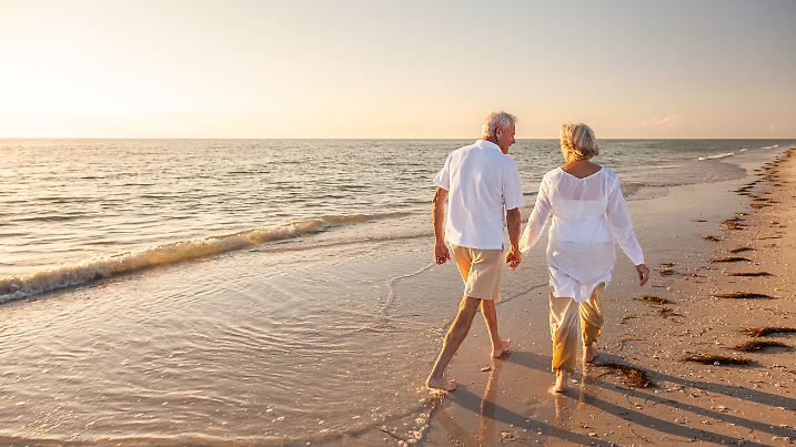 Glückliches älteres altes pensioniertes Paar, das am Strand bei Sonnenuntergang spazieren geht und sich an den Händen hält