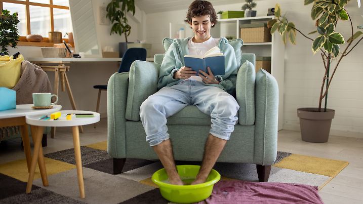 A young Caucasian man having a foot bath and reading in the living room of his apartment