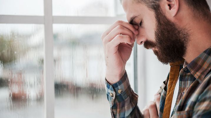 Depressed young bearded man keeping eyes closed and touching his face while standing near the window