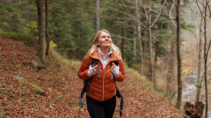 A happy woman enjoys a refreshing hike in an autumn forest, wearing a puffy orange jacket and carrying a backpack. The autumn foliage provides a picturesque and serene natural backdrop.