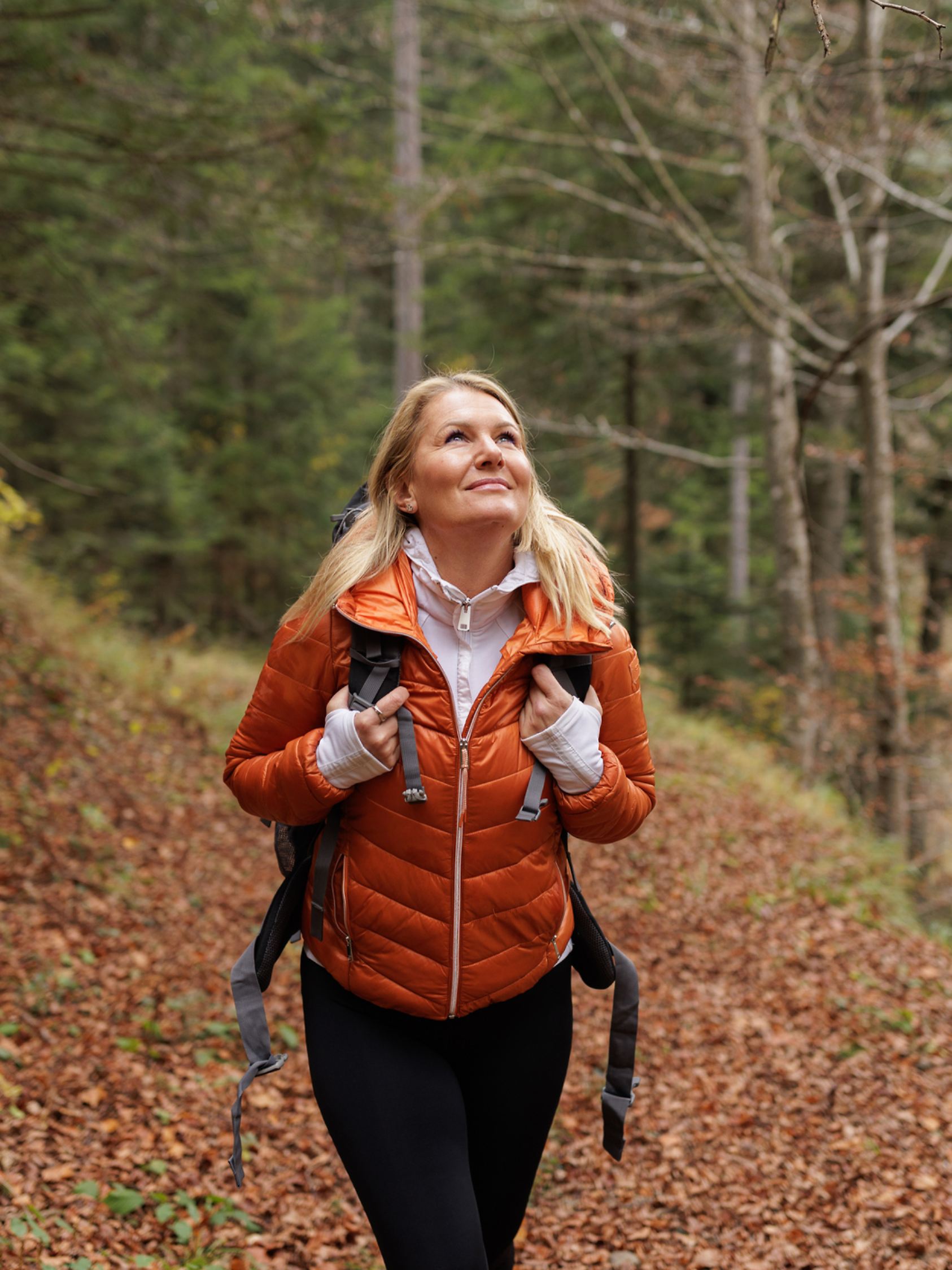 
A happy woman enjoys a refreshing hike in an autumn forest, wearing a puffy orange jacket and carrying a backpack. The autumn foliage provides a picturesque and serene natural backdrop.