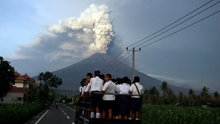 dpatopbilder - Schüler fahren am 28.11.2017 in Karangasem, Bali, Indonesien, auf ihrem Weg zur Schule auf einen Lastwagen, während der Vulkan Mount Agung im Hintergrund Rauch und Asche spuckt. Auf der indonesischen Ferieninsel Bali wird ein gewaltiger Ausbruch des Mount Agung befürchtet. Nach einer Reihe von kleineren Eruptionen gilt seit dem 27.11.2017 rund um den mehr als 3000 Meter hohen Berg Alarmstufe Rot. Die indonesischen Behörden forderten 100 000 Menschen auf, sich in Sicherheit zu bringen. Beim letzten großen Ausbruch des Gunung Agung - wie der Vulkan in der Landessprache heißt - hatte es mehr als 1100 Tote gegeben. Foto: Firdia Lisnawati/AP/dpa +++(c) dpa - Bildfunk+++