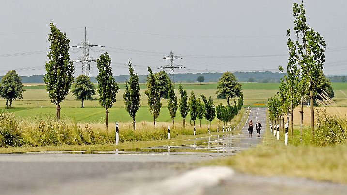 Zwei Frauen wandern am 27.06.2017 südlich von Hannover (Niedersachsen) auf einer Straße. Dabei führt die Hitze über dem Asphalt zu Luftspiegelungen. Solch eine Fata Morgana sieht aus, als würde Wasser auf der Straße stehen. Foto: Holger Hollemann/dpa | Verwendung weltweit