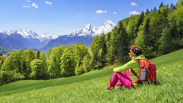 Frühlingshafte Hügellandschaft oberhalb von Berchtesgaden unterhalb der Kneifelspitze im Frühjahr vor den noch tief verschneiten Bergen der Berchtestgadener Alpen mit dem Watzmann. Eine Frau sitzt in der Wiese und genießt den wunderbaren Tag, die frische Luft und die herrliche Aussicht - 20190502_PD12491 |